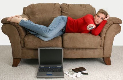 Beautiful young woman taking a break from balancing her bills online. Laptop, cash, credit card, bills and checkbook on the floor in front of her as she relaxes on the couch watching tv.